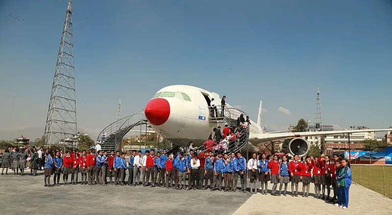 A white airliner with a red nose parked on the grass, with a large school group lined up in front of it