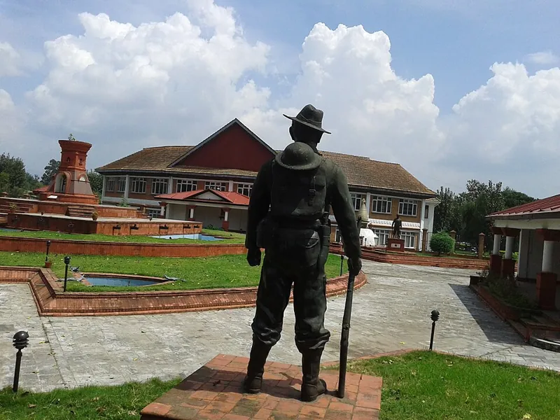 A bronze statue of a soldier in a wide-brimmed hat, looking across a garden toward the museum building