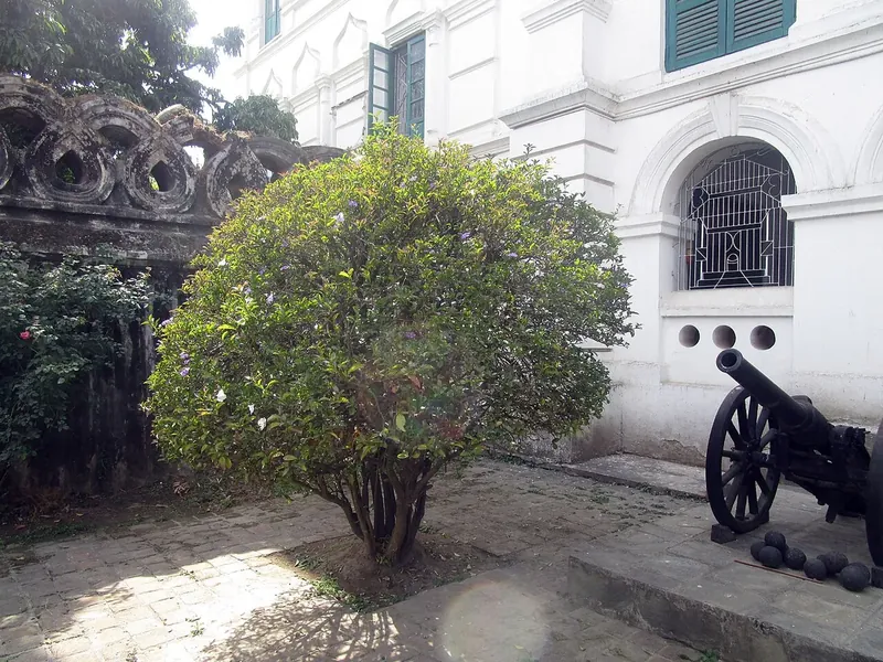 An old cannon on a paved courtyard beside a white building with green shutters