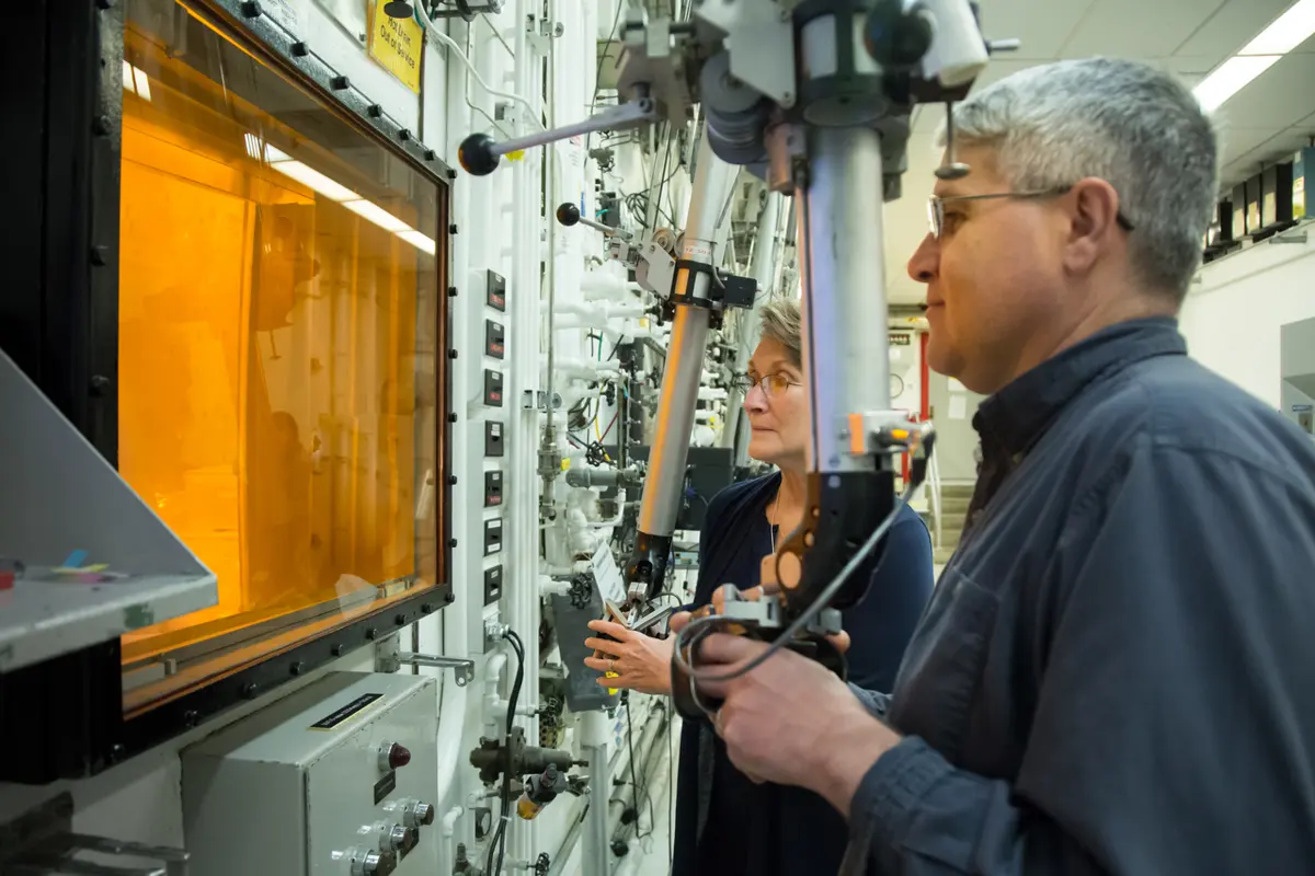 A man and a woman in a laboratory working long metal handles that reach through the wall of a shielded cell, looking through a thick orange-tinted window