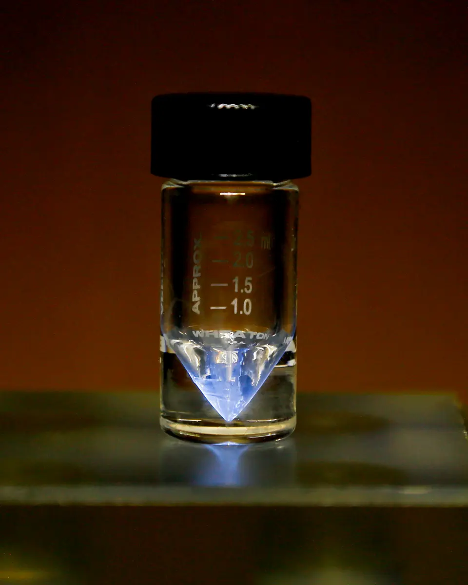 A small glass vial with a black cap and measuring marks, its pointed bottom glowing blue, on a glass shelf in a dim room