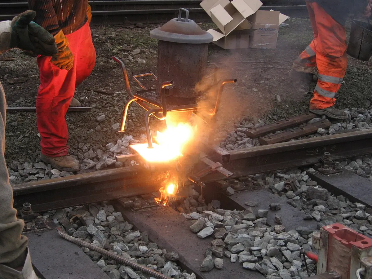 Workers in orange trousers beside a railway track; a dazzling orange glow and smoke pour from a small pot set over the gap between two rails