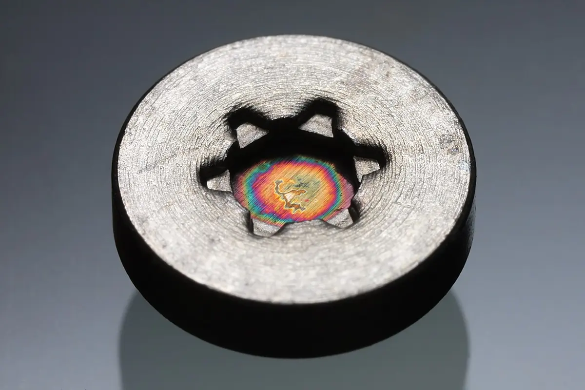 A small round gray metal button, about the size of a shirt button, with a six-pointed slot in its face through which a shiny spot with rainbow colors shows