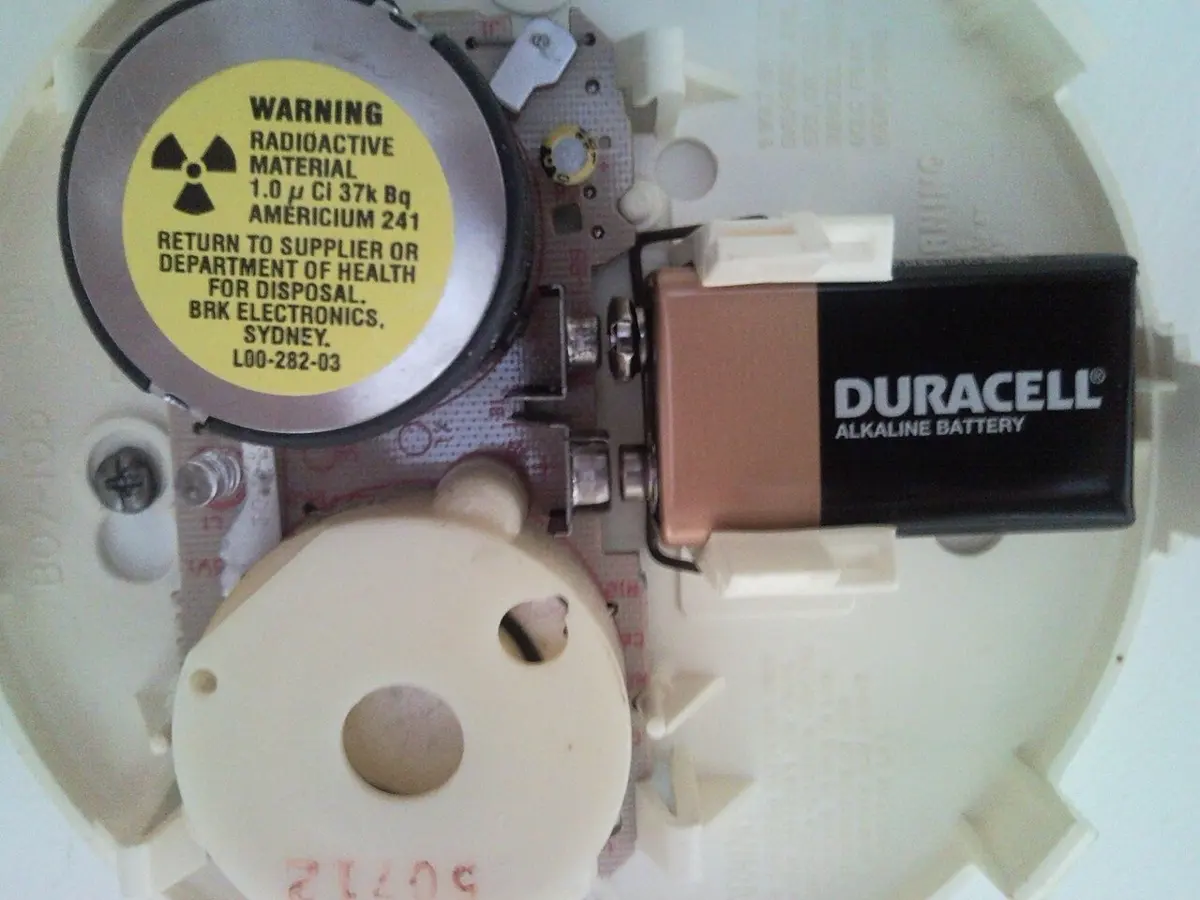 The inside of a smoke alarm with its cover off: a round metal chamber with a yellow warning label with the radiation sign and the words Radioactive material, Americium 241, beside a 9-volt battery