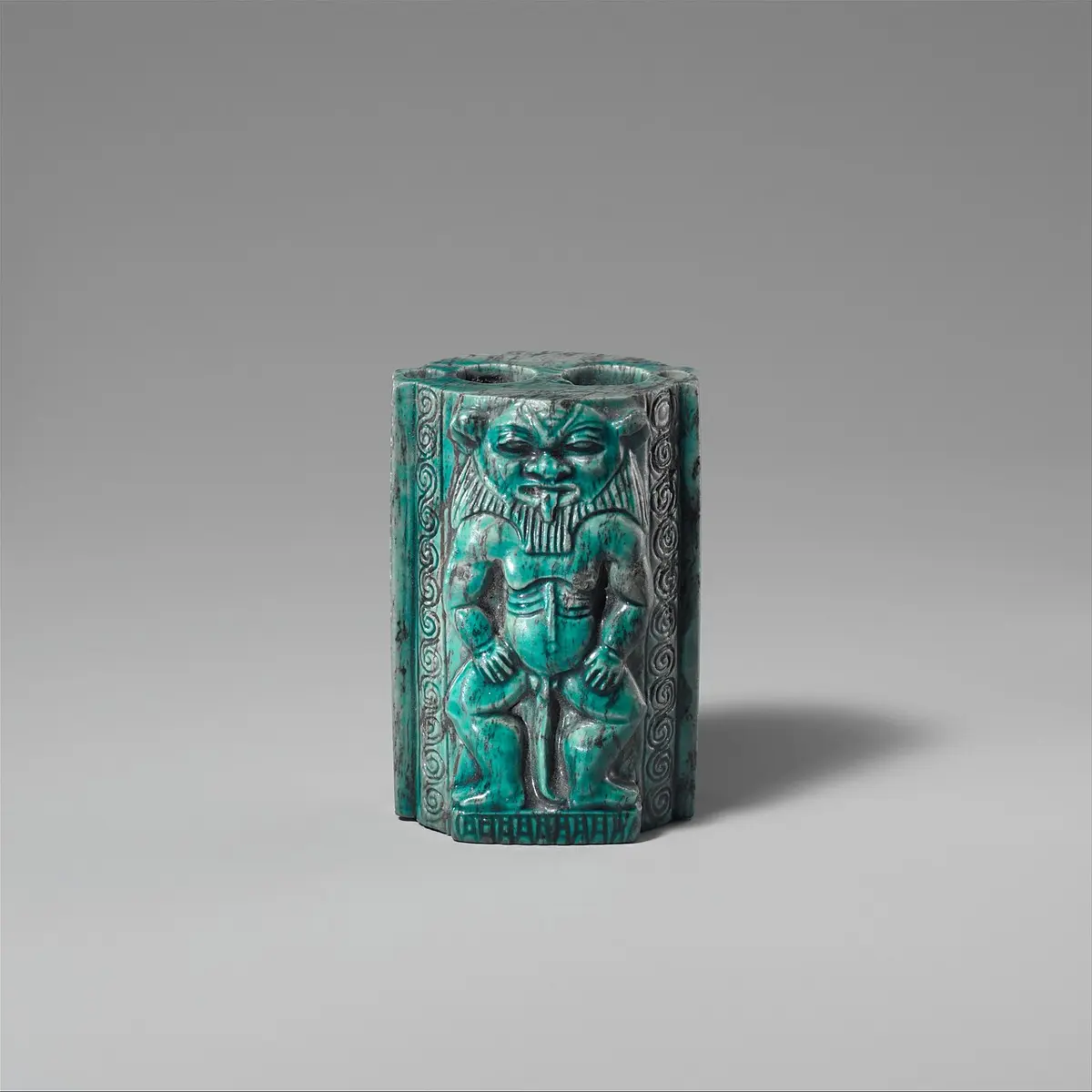 A small blue-green stone container with two round holes in its top, carved on the front with a squat bearded figure standing between bands of spirals