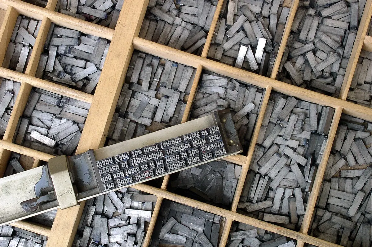 A wooden tray divided into compartments full of small gray metal blocks, with a metal holder on top holding a line of blocks whose ends carry raised, back-to-front letters