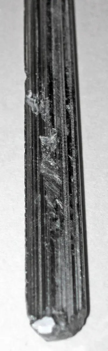 A long, straight, dark gray crystal with fine grooves running along its length, shiny in places, on a white background