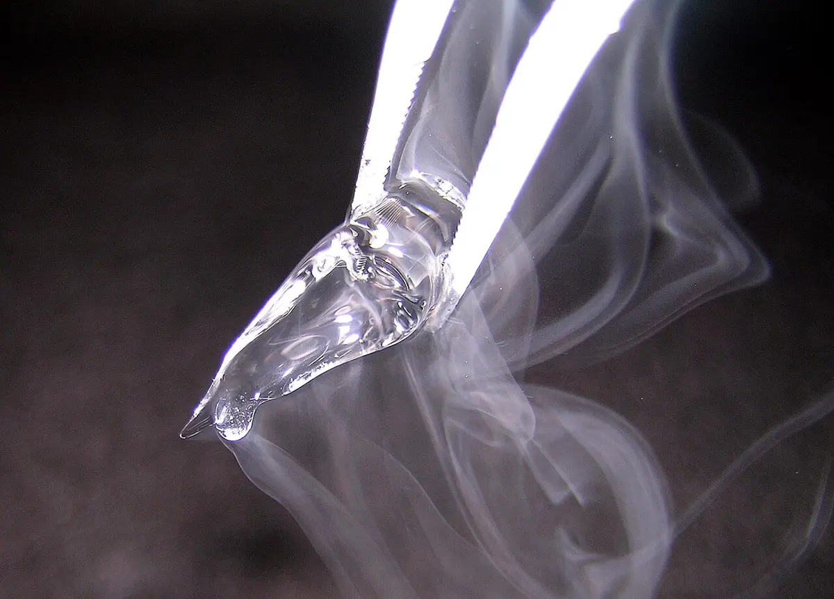 A small clear lump of solid held in metal tweezers, with white mist streaming off it