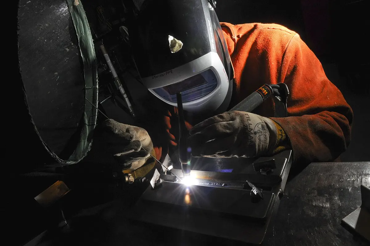 A welder in a dark helmet, orange jacket and gloves bending over a metal part, with a bright white spark at the tip of the torch