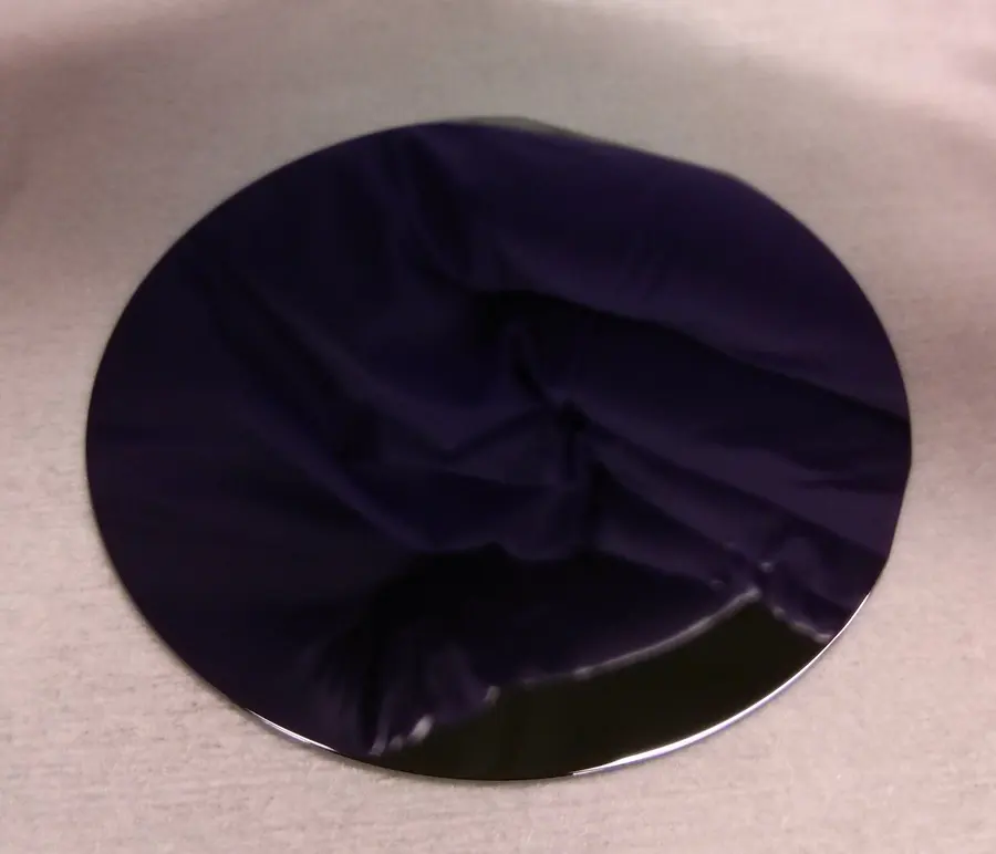 A thin, round, very dark disc with a mirror-like surface, lying on a pale surface; a purple reflection shows in it