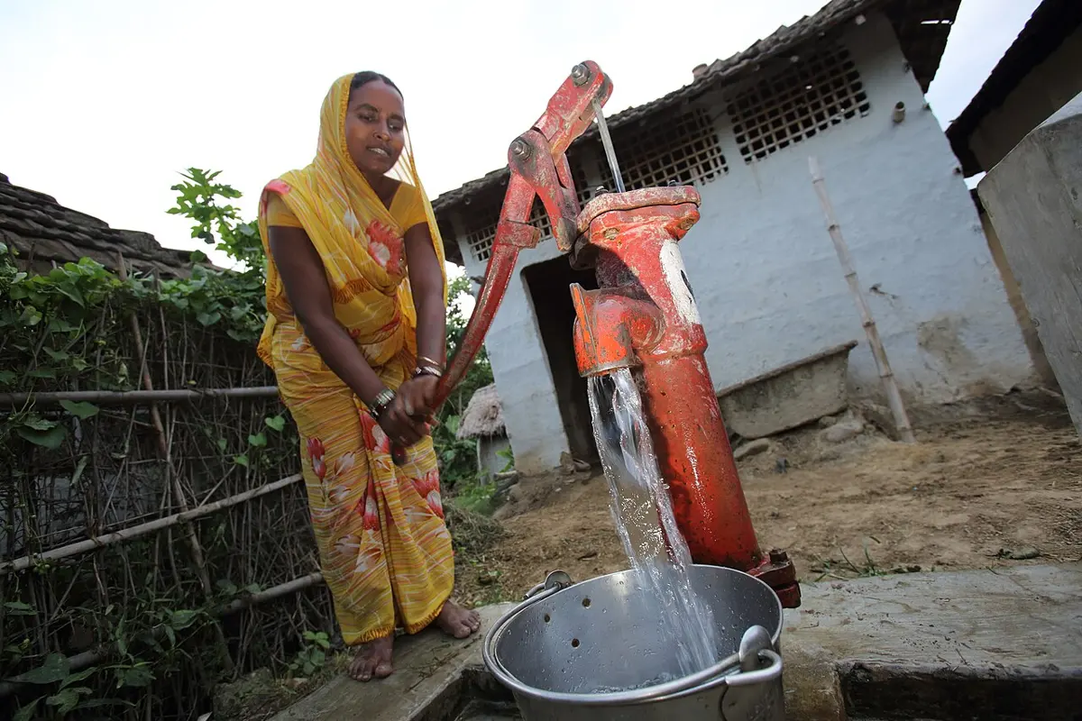 A woman in a yellow sari pumps the long red handle of a hand-pump tube well, filling a steel pot with water, outside a village house