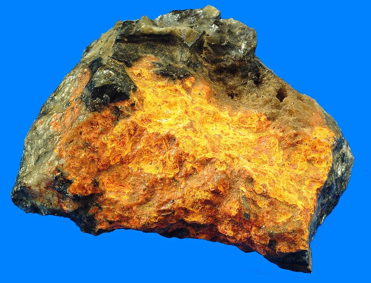 A rough lump of rock, mostly bright orange-yellow, with darker grayish patches on top, photographed against a blue background
