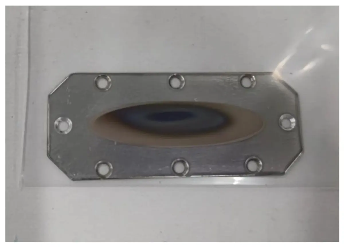 A flat, eight-sided metal plate with screw holes round its edge and an oval brown and blue patch in the middle