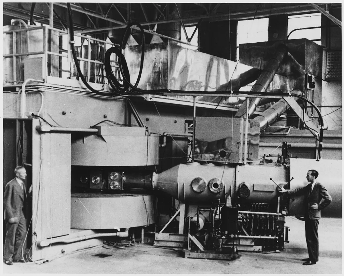 A black and white photograph of a large machine in a hall: two huge round magnet coils on the left, a long metal tank with round windows and pipes on the right, and a man in a suit standing at each end