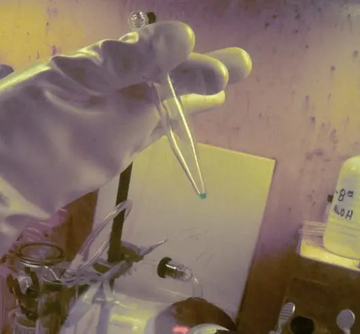 A gloved hand inside a sealed glass box holds a narrow plastic tube; a tiny green drop sits at its very tip