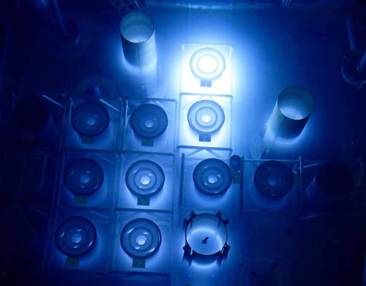 Looking down through deep water at rows of round, numbered metal cylinders standing in racks, all lit by a bright blue glow