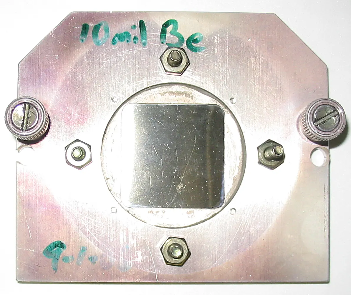 A square of shiny metal foil in a round opening in a metal plate held by screws, with “10 mil Be” written on the plate in green ink