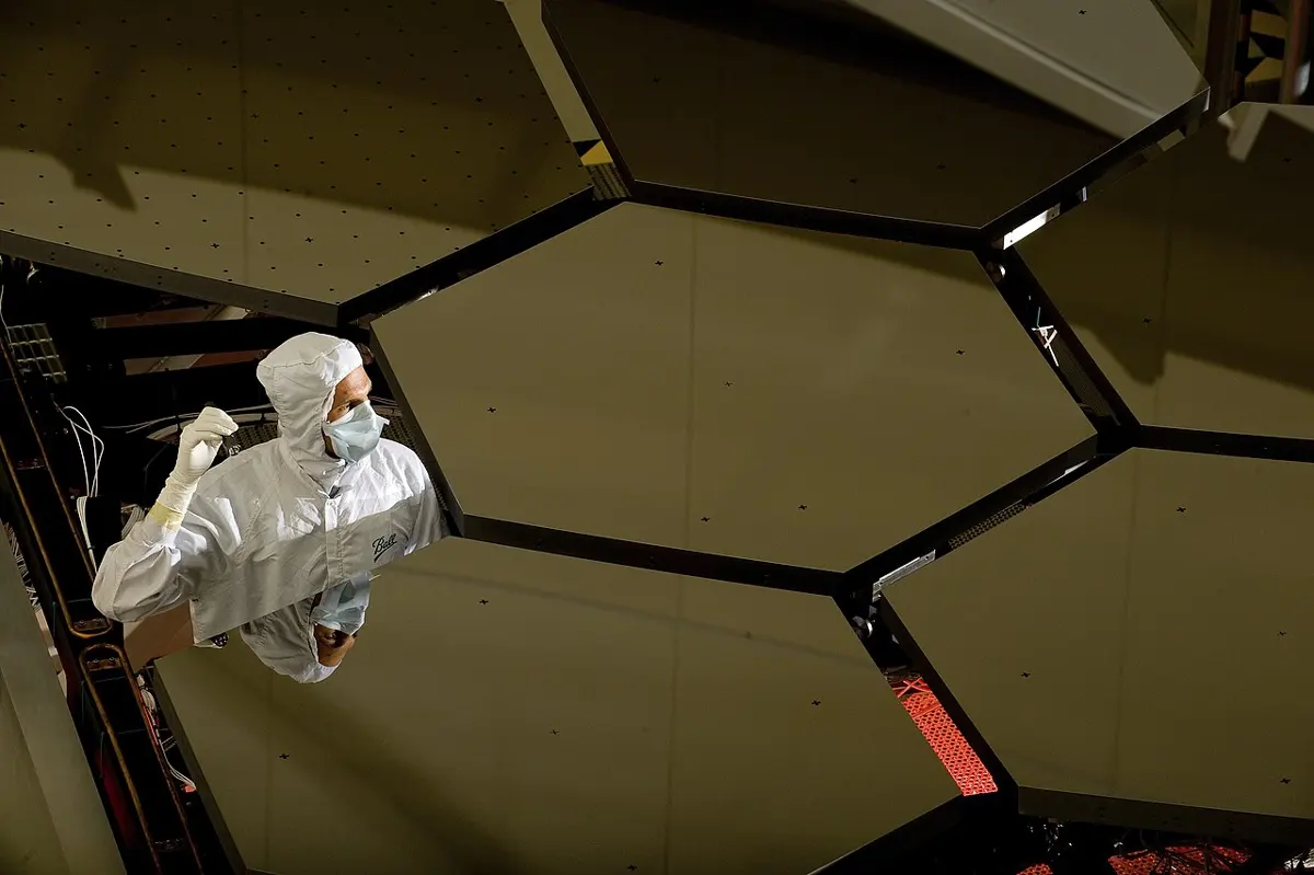 Six large six-sided mirror pieces fitted side by side, seen from below, with a worker in a white clean-room suit, mask and gloves reflected in them