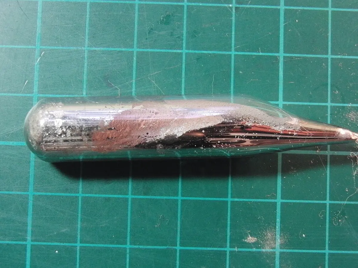 A sealed glass tube on a green squared cutting mat, holding shiny silvery metal with a faint pink sheen