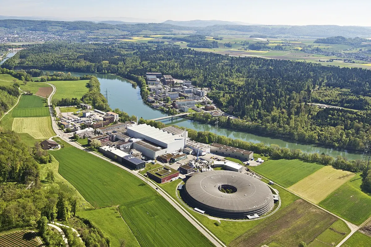 An aerial photo of a river winding between green fields and woods, with laboratory buildings on both banks and a large round building on the near side