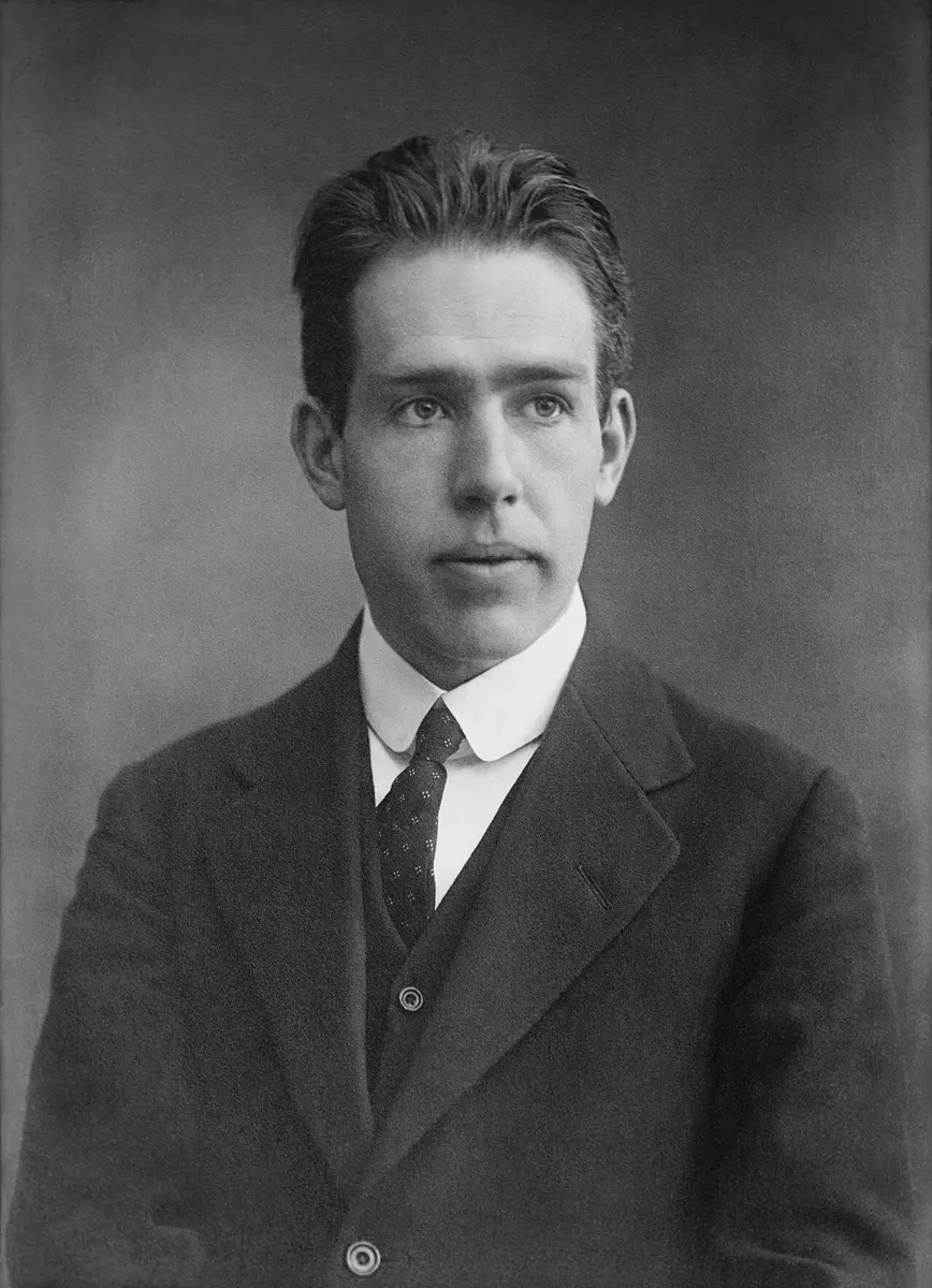A black-and-white portrait of a young man with dark, swept-back hair, in a dark suit, waistcoat and tie, looking towards the camera