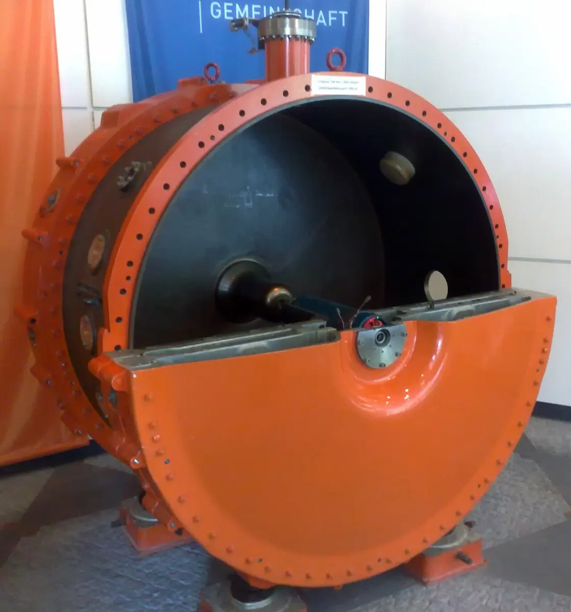 A large orange metal cylinder, open at the front like a drum, with a copper tube and parts inside, standing on a stand in a hall