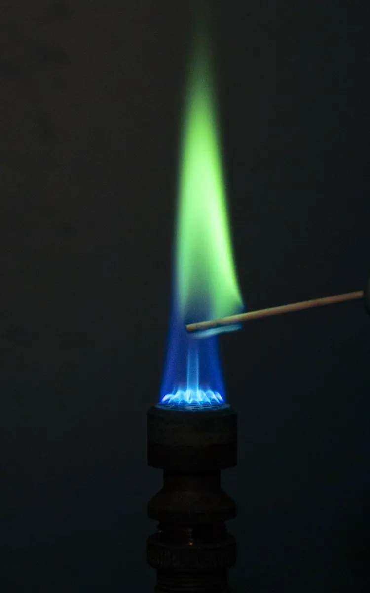 A gas burner with a blue flame at its base and a tall bright green flame above it, beside a thin wooden splint