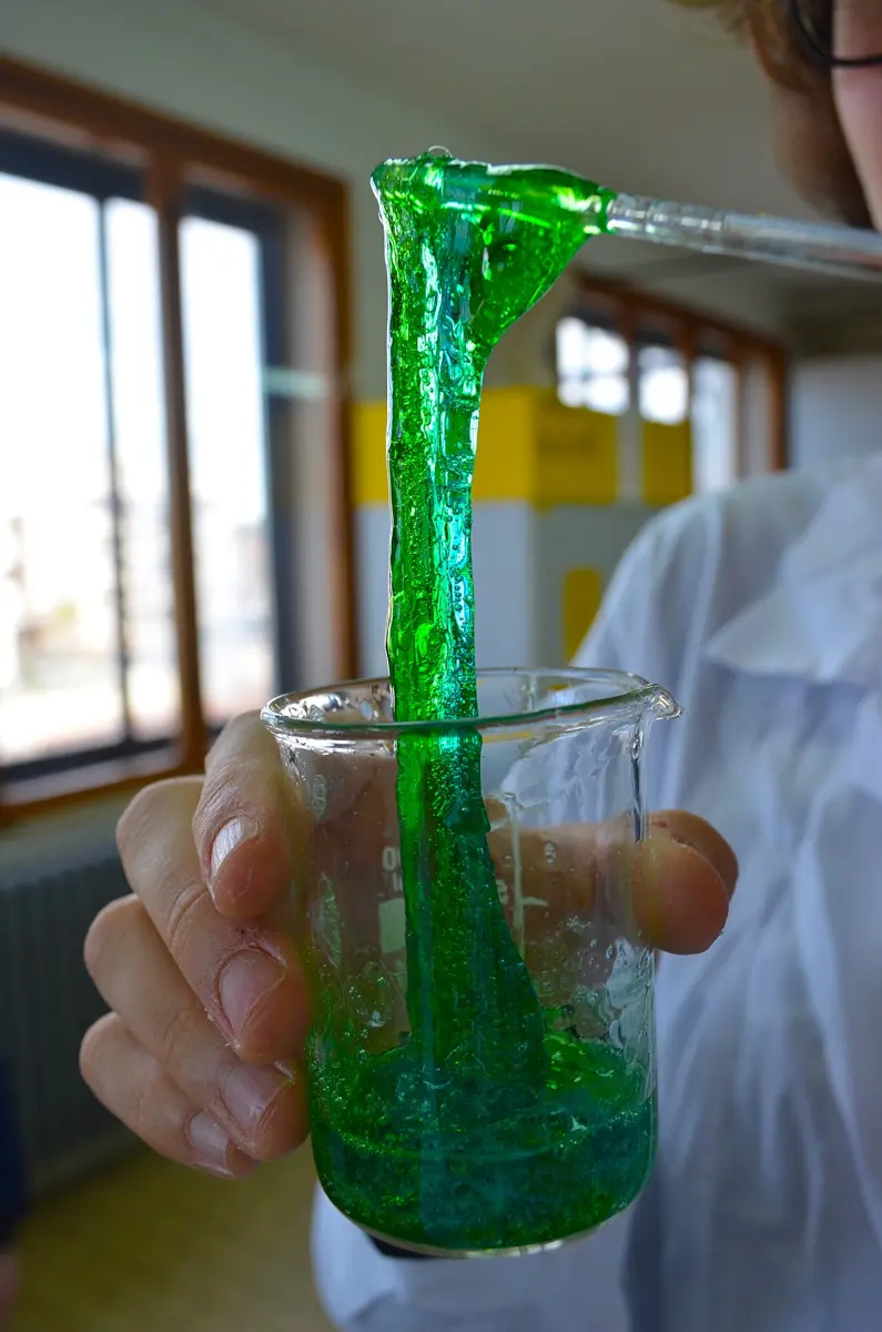 A hand holding a glass beaker of bright green slime, with a long, drooping string of it pulled up out of the beaker on a glass rod