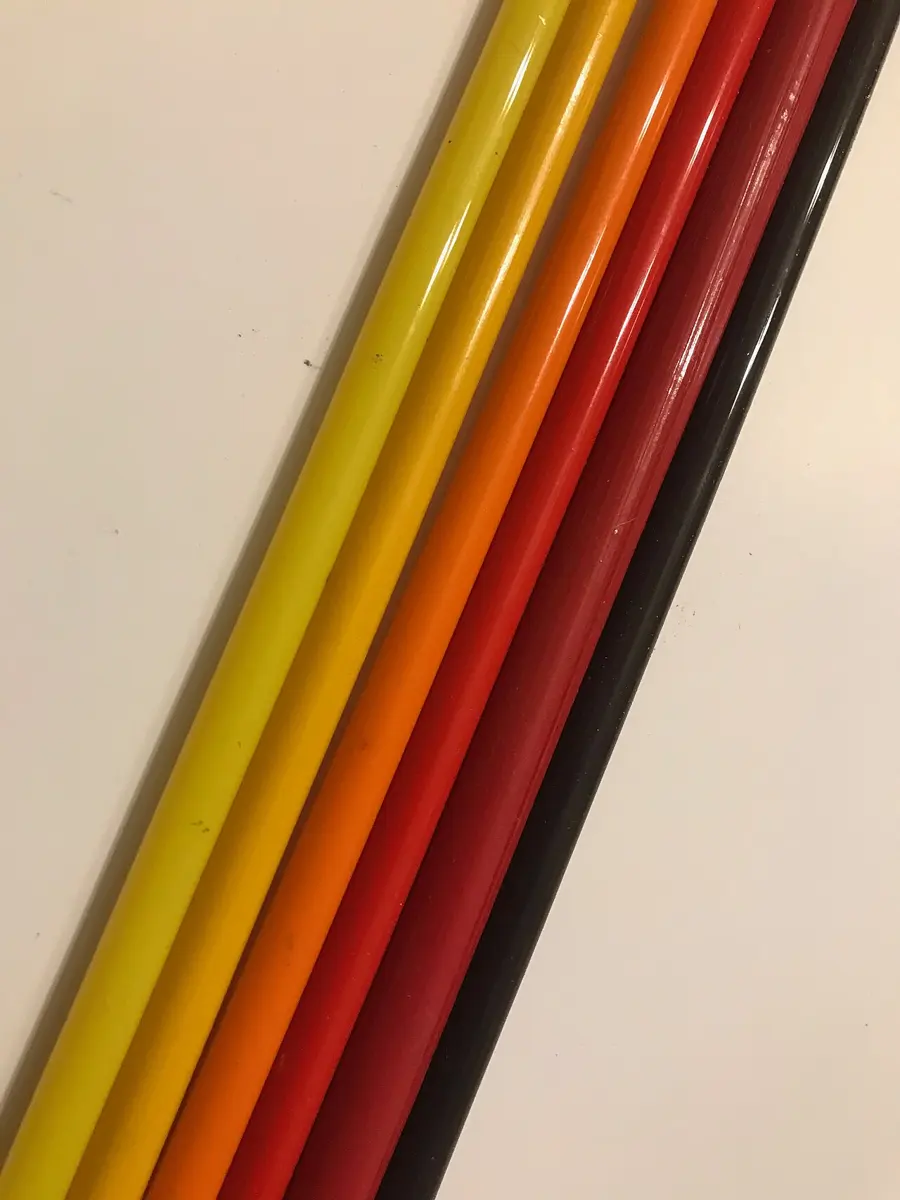 Six shiny glass rods lying side by side on a white surface, colored lemon yellow, golden yellow, orange, red, dark red and almost black