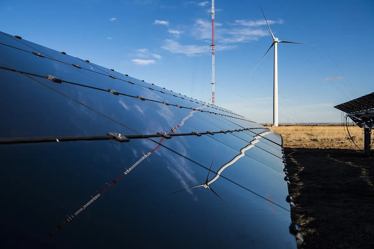 A long row of smooth, dark blue solar panels under a blue sky, reflecting a wind turbine and a tall red-and-white mast that stand behind them in a dry field