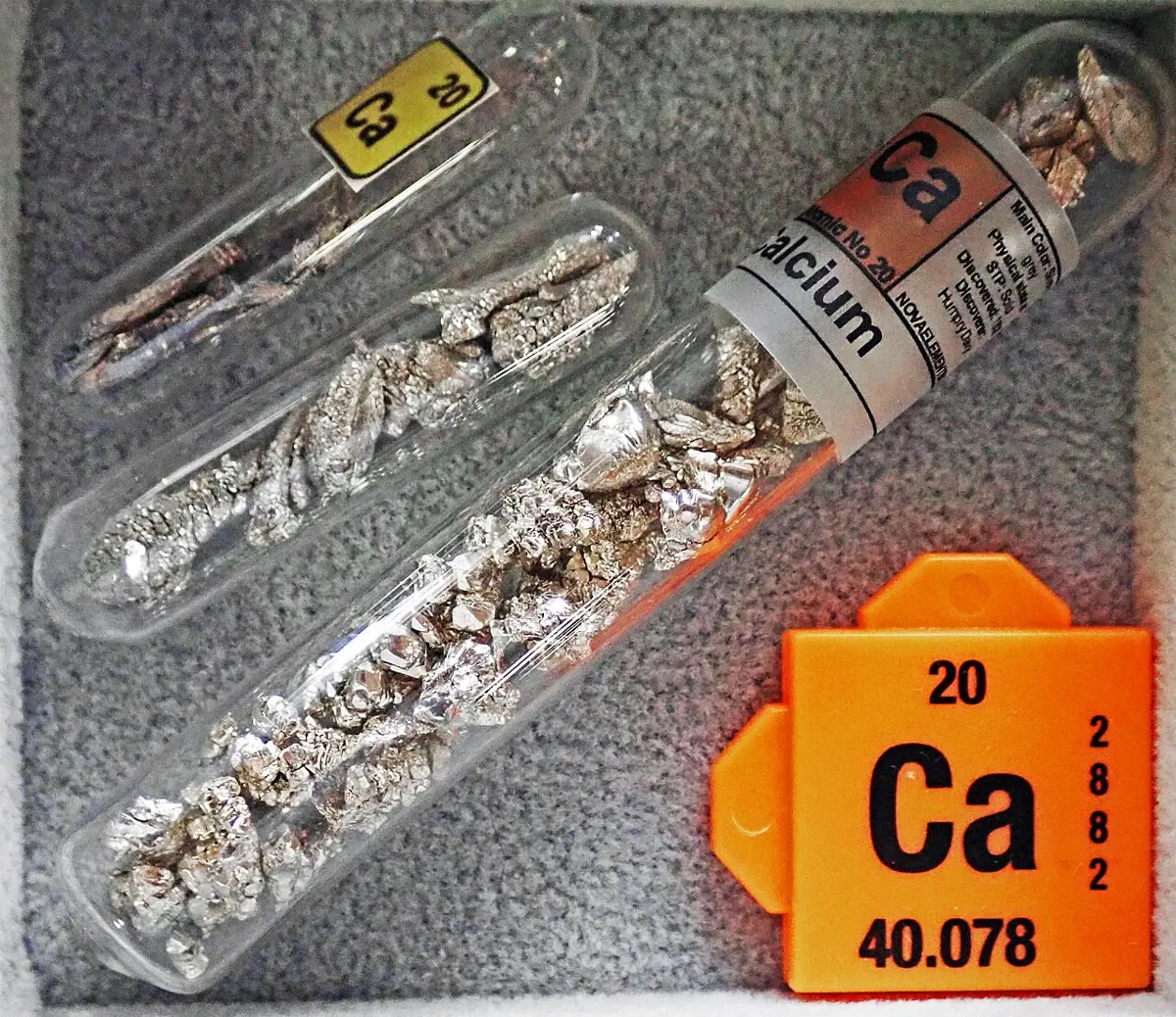 Three sealed glass tubes of silvery, crinkled flakes and chunks of metal, beside an orange tile reading 20, Ca, 40.078