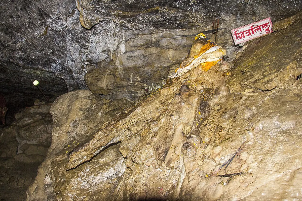 Inside a dim cave: pale, rounded and dripping rock formations under a dark rock roof, with a red sign in Devanagari and an orange cloth tied on one formation