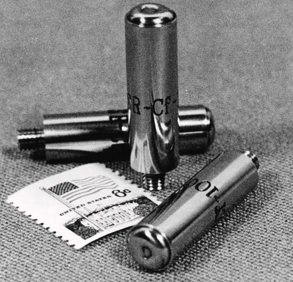 A black-and-white photo of three shiny metal capsules, each a few centimeters long and marked SR-Cf-100, lying beside a United States postage stamp