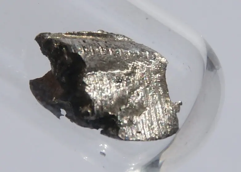 A rough, silvery-gray lump of metal with a darkened side, in a clear glass tube
