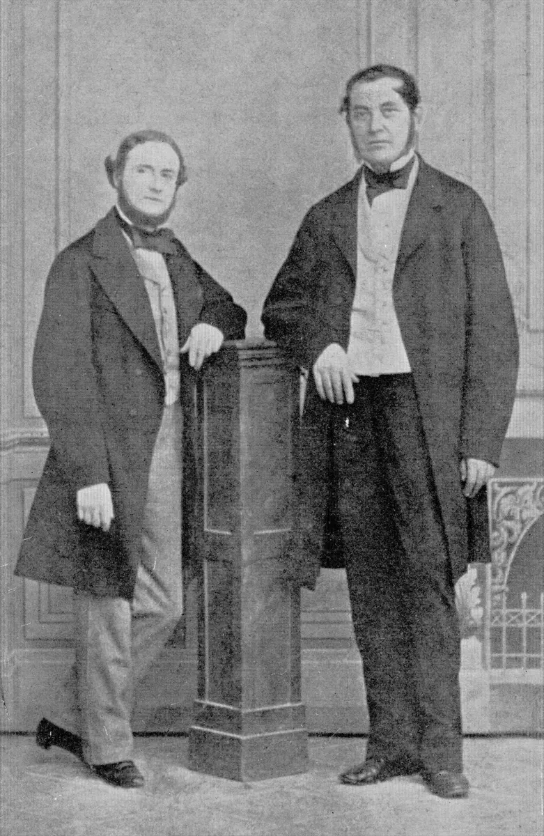 An old black-and-white photograph of two men in long dark coats, standing beside a tall pedestal; the man on the left leans on it