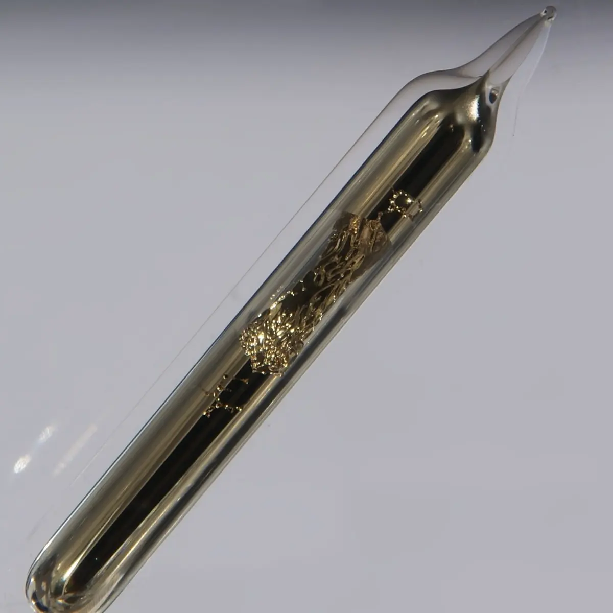 A sealed glass tube, pointed at the top, lying at a slant, with shiny, golden crystals of metal spread along the middle