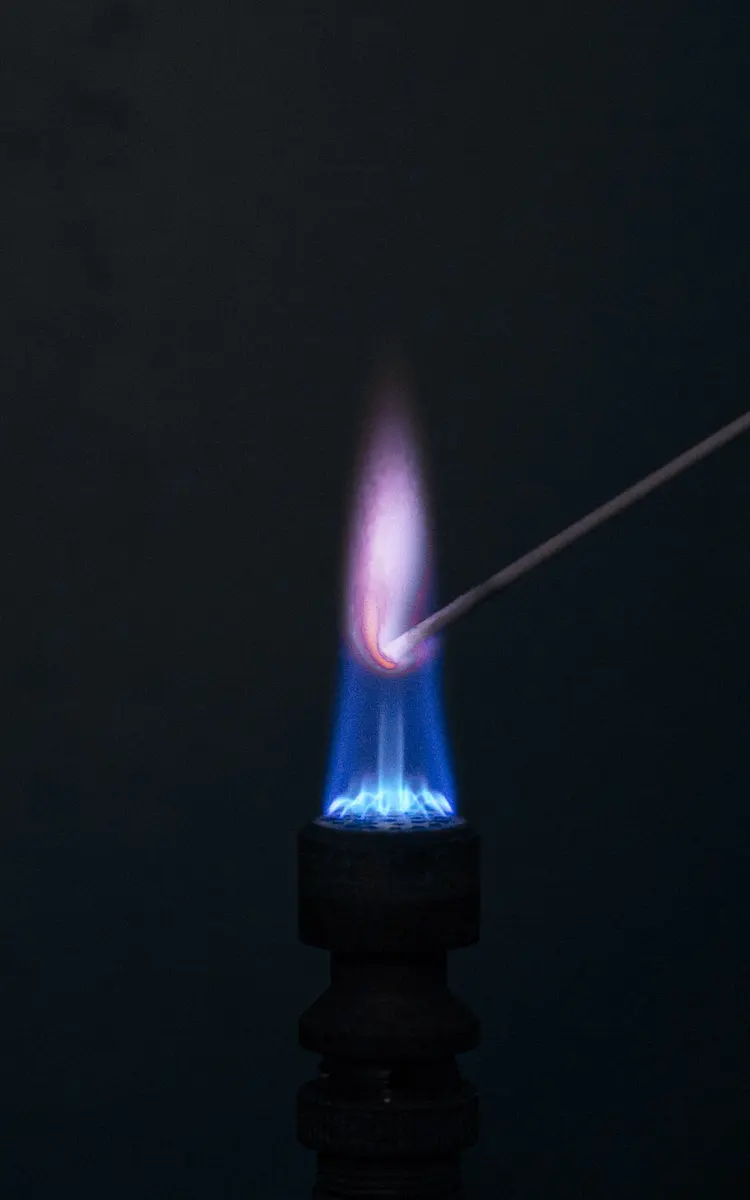 A blue gas flame on a dark background with a thin wire held in it; above the wire the flame is a pale violet-pink