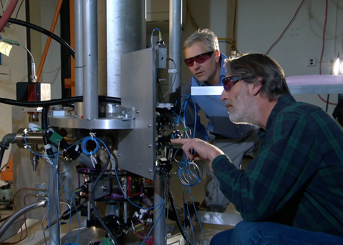 Two men in safety glasses working at a tall metal apparatus full of pipes, cables and small boxes, in a laboratory