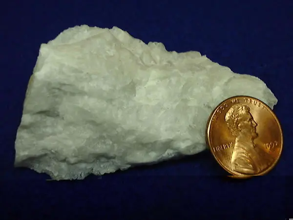 A rough lump of pale, whitish mineral beside a copper one-cent coin, on a dark blue background