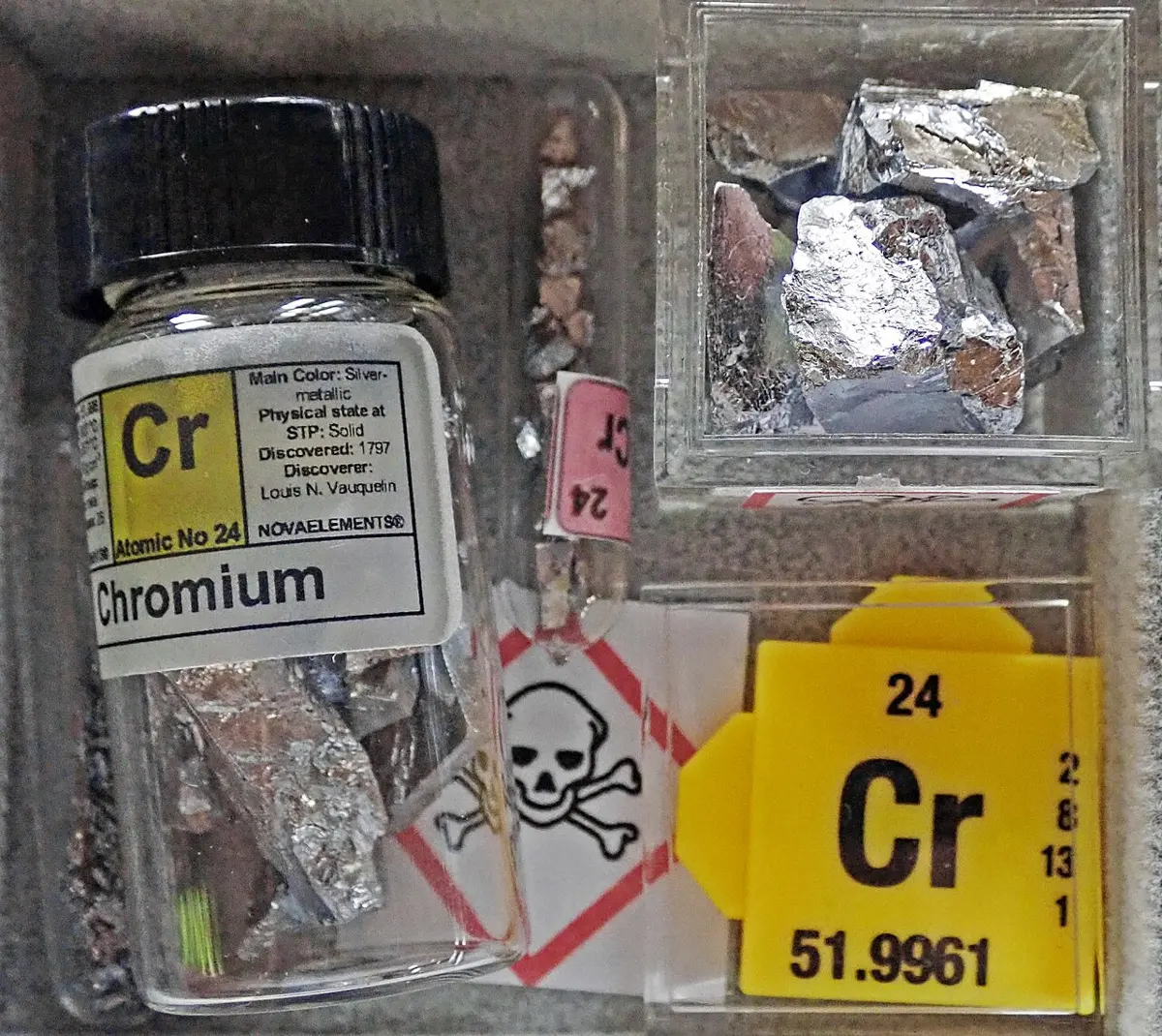 Shiny, silvery-gray flakes and lumps of metal in a glass jar labeled Chromium and in a small clear box, beside a yellow card that reads Cr, 24 and 51.9961