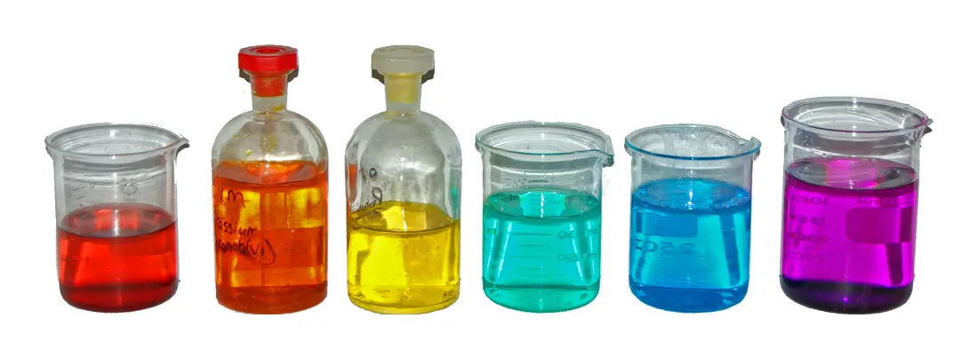 Six glass beakers and bottles in a row, holding clear solutions colored red, orange, yellow, turquoise, blue and purple