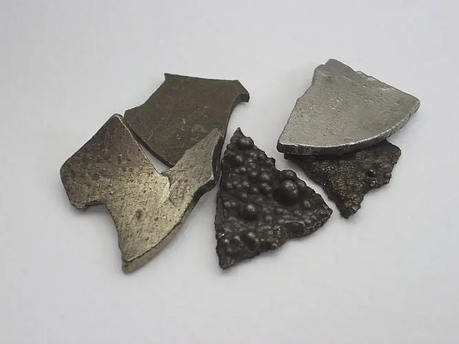 Six flat, broken pieces of gray metal on a white surface, some smooth and shiny, two dark and covered in small round bumps