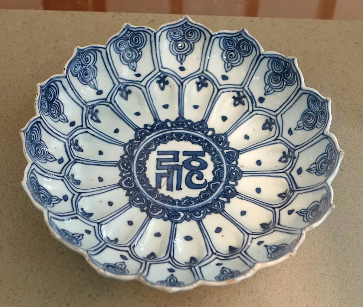A white porcelain dish shaped like an open flower with many petals, painted in deep blue, with a letter of an Indian script in a circle at its center