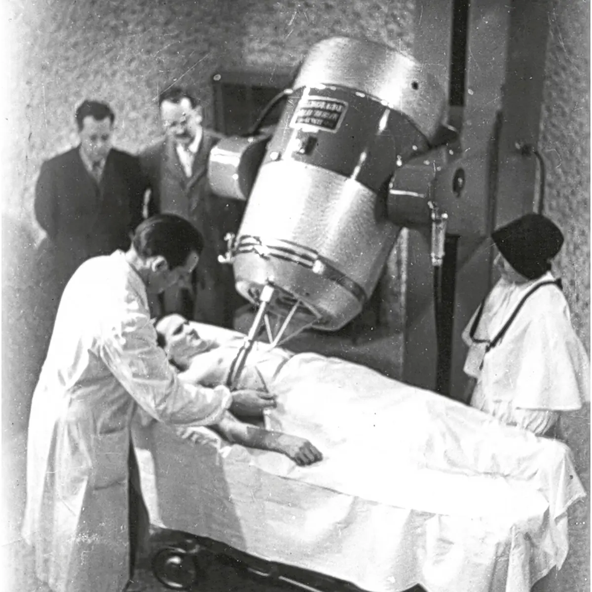 An old black-and-white photo: a doctor in a white coat bends over a patient lying on a table under a large round machine head, watched by two men in suits and a nun