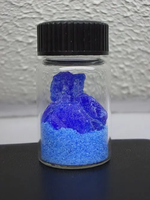 A small glass jar with a black lid, holding dark blue crystals on top of light blue powder