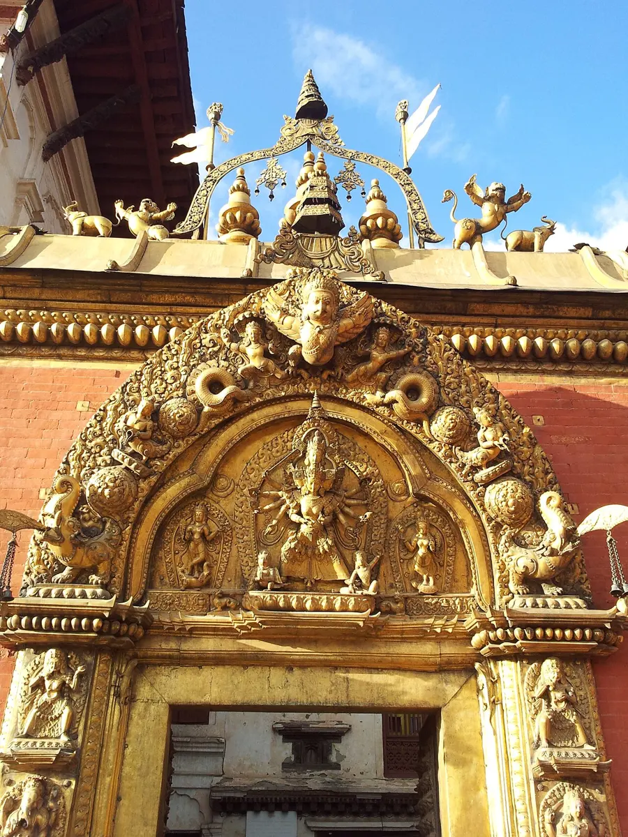 An ornate golden doorway in a red brick wall, with a carved arch of gods, a winged Garuda at the top and a many-armed goddess in the middle