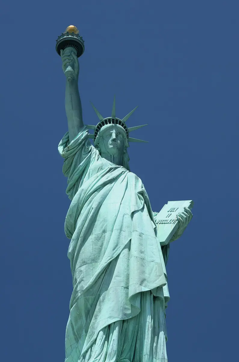 The Statue of Liberty from the front: a pale green robed figure holding up a torch, standing on a tall stone base under a blue sky