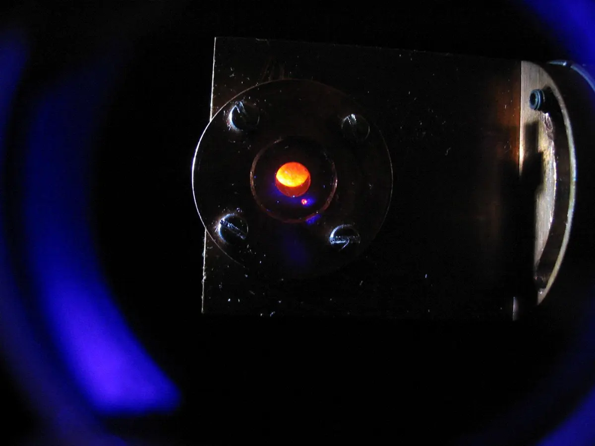 A dark photo of a metal holder with a small window, in which a bright orange spot glows, with dim blue light around it