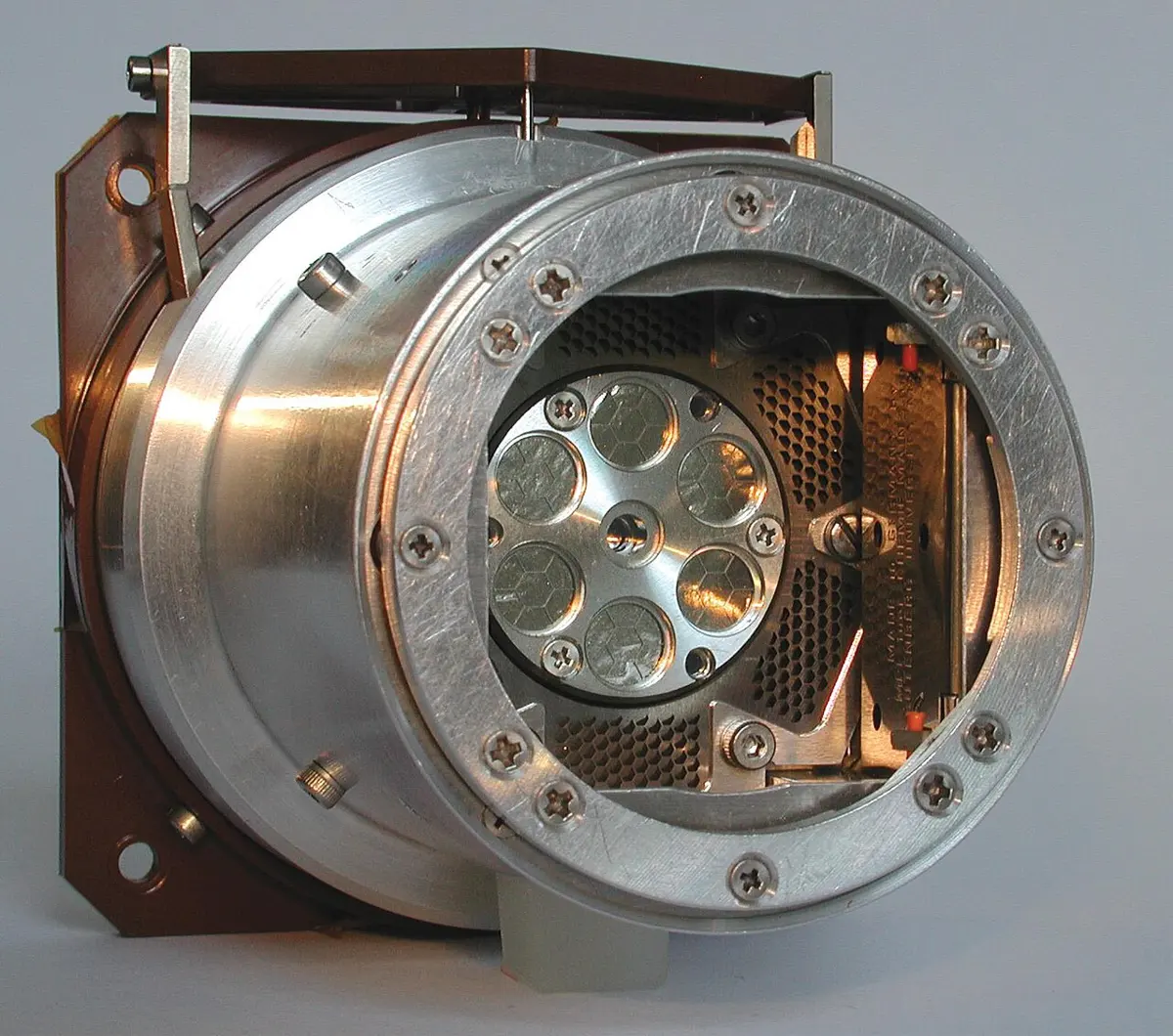 A shiny metal cylinder seen from the front at an angle; inside its open end, six small round discs in a ring behind a fine mesh