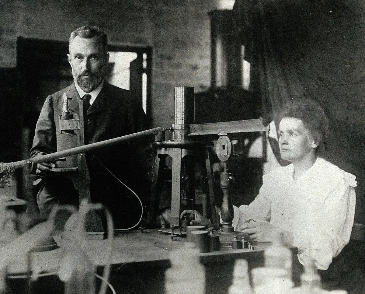 A black-and-white photo of a man and a woman in a laboratory, standing beside a table of scientific apparatus; the woman is working the apparatus